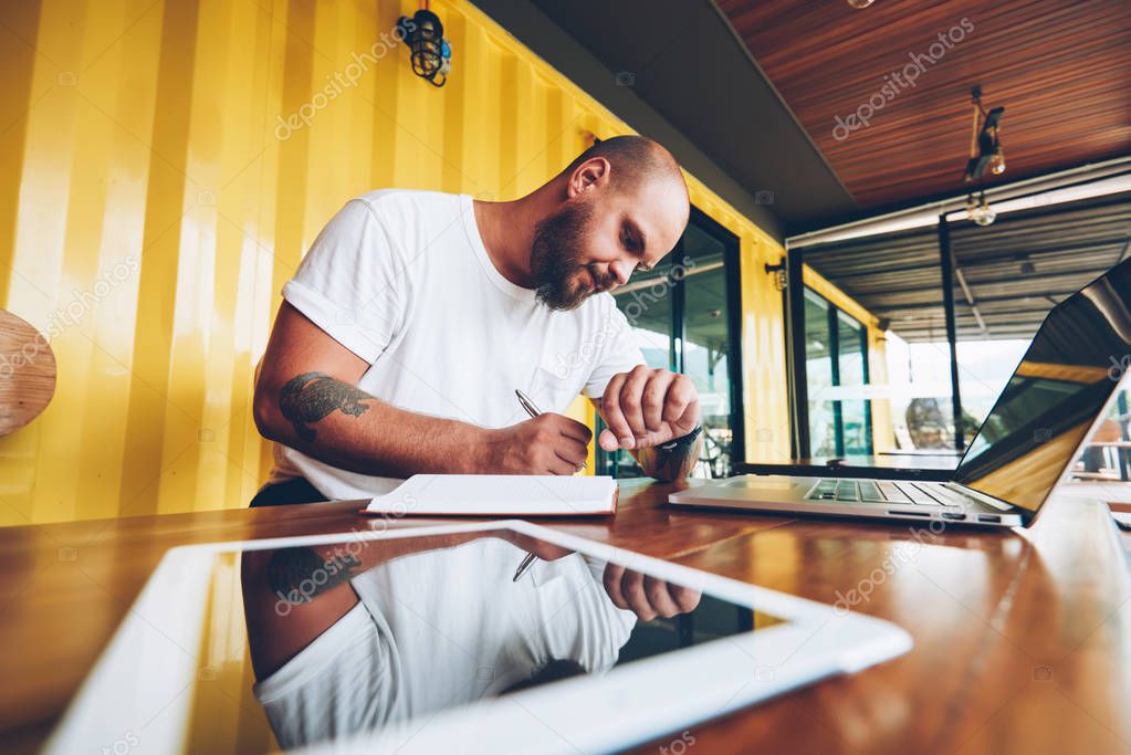 Pensive male freelancer making notes and managing time on wrist watch sitting near laptop computer for job, serious young man looking at time planning working day and writing ideas in notebook