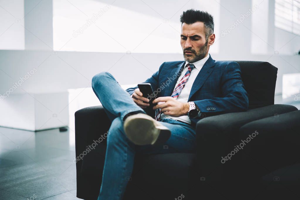 Confident businessman in formal wear watching video online on website on smartphone resting in office on armchair.Pensive executive director 50 years old reading incoming notification on telephone