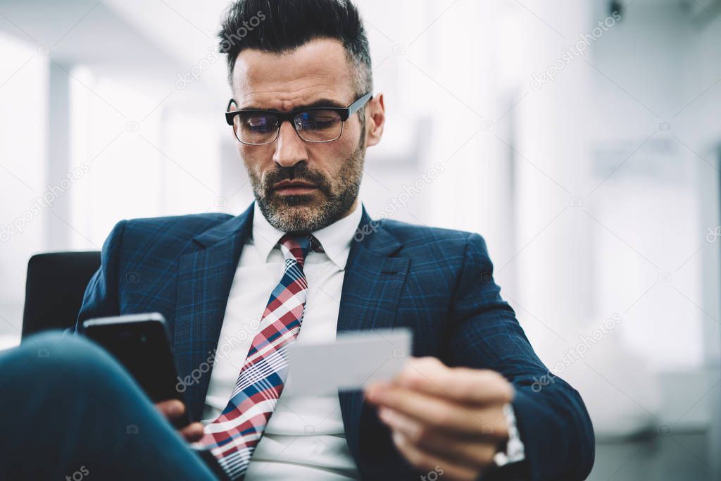 Confident mature executive manager in formal wear dialing phone number from business card on smartphone device.Serious entrepreneur 50 years old reading notification on modern telephone