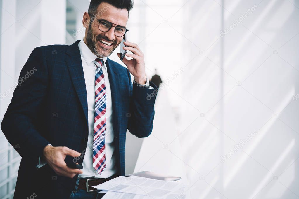 Portrait of successful businessman dressed in formal wear calling on smartphone device and laughing during phone conversation standing in office of corporate company near copy space area