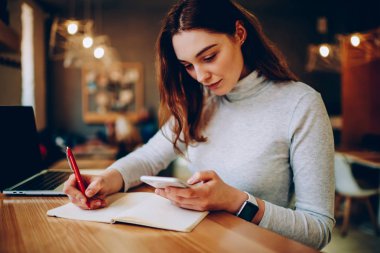 Uzaktan coworking alanda, bilgi aramak için teknoloji ve kablosuz bağlantı kullanarak ödev genç kadın çalışma Not defteri cep telefonu kişiden belirterek ciddi kadın serbest meslek