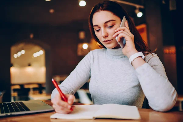 Serious woman freelancer making notes during business conversation on telephone working distanly in cafe, pensive female noting information calling for banking customer support for consultancy