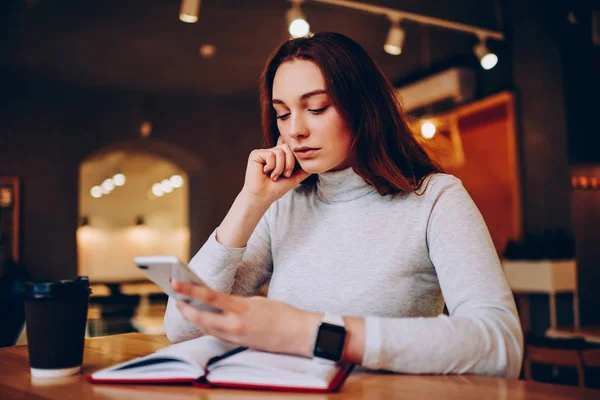 Serious female student reading news on web page of college browsed on cellphone while learning in cafeteria, pensive young woman checking mail with receipts for paying in application on telephone