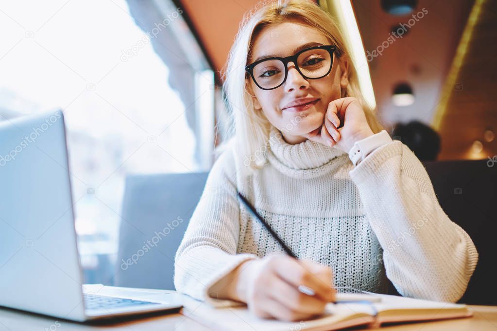 Portrait of skilled woman in spectacles planning working process for startup making notes satisfied with freelance business, clever female student writing in notepad doing homework in cafe interio