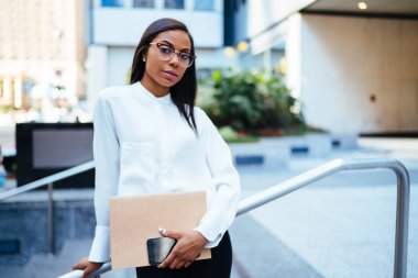 Kendine güvenen kadın ofis çalışanı ile karanlık yarım uzunluk portre fotoğraf makinesi ayakta kentsel ortamda bakarak derili. Ciddi Afro-Amerikan işkadını klasör ve akıllı telefon elinde tutan