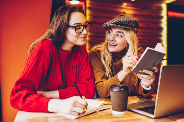 Happy female students cooperating for completing common project learning in cafe interior with laptop computer,cheerful girls discussing creative ideas for freelance startup in friendly atmosphere