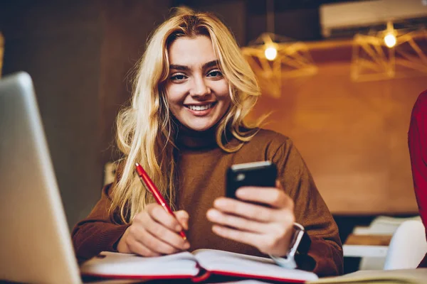 Portrait of cheerful hipster girl writing in notepad and holding smartphone for chatting, successful female blogger editing text of article in notebook before post on social networks on telephon