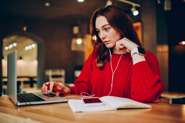 Pensive hipster girl preparing for exams searching information on netbook and listening music on cellphone for concentration,clever female student learning language via apps on smartphone and lapto