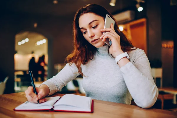 Serious young woman writing information in notebook while talking on telephone in coworking space,concentrated female calling for consultancy to helpline operator making notes while communicating