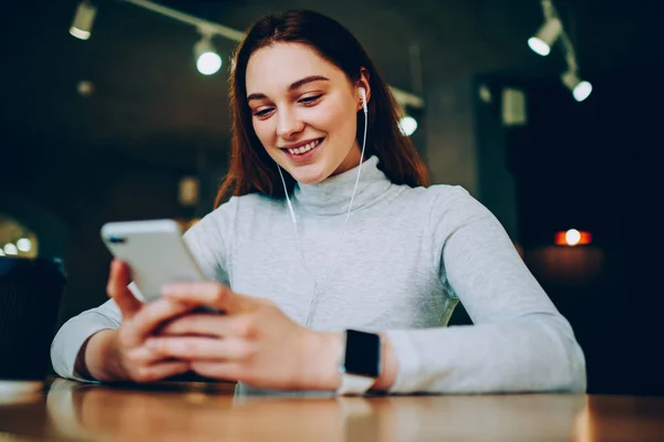 Cheerful teen girl watching favorite serial on smartphone via good wifi connection in cafe, smiling young woman using cellular for downloading new songs for playlist listening music on coffee brea