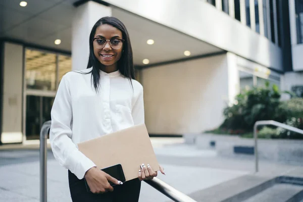 Half length portrait of prosperous black skinned employee in formal wear smiling at camera while holding folder and smartphone.Successful african american businesswoman standing near office building