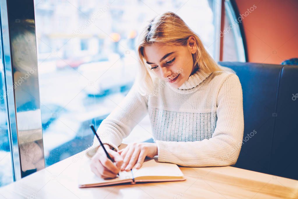 Clever blonde female student writing in notepad doing college homework task spending leisure time  in cafe interior,creative girl blogger making notes of ideas for article publication in notepad