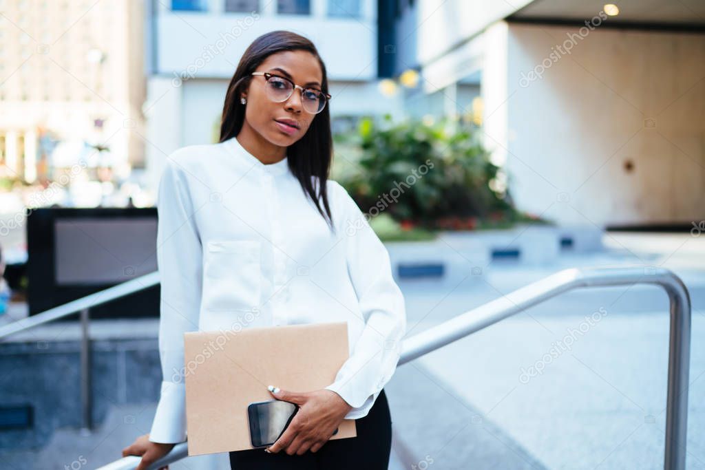 Half length portrait of confident female office employee with dark skinned looking at camera standing in urban setting.Serious african american businesswoman holding folder and smartphone in hands