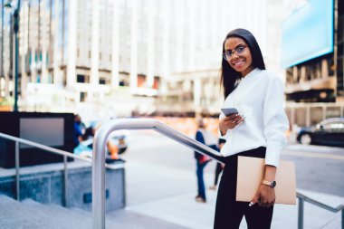 Klasör çevrimiçi smartphone aygıt ayakta şehir merkezinde sohbet elinde başarılı koyu tenli avukatla tam uzunlukta portresi. Müreffeh Afrikalı-Amerikalı genç kadın üzerinde cep SMS gönderme