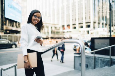Smartphone'da bankacılık uygulaması yüklenirken kameraya gülümseyen klasörle Afro-Amerikan başarılı erkek portresi. Neşeli ofis çalışanı beyaz gömlekli profil sayfası telefon ayakta City güncelleştiriliyor