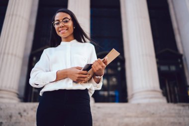 Aşağıda görünümü kamera ayakta university yakınındaki gülümseyen koyu tenli neşeli kadın avukat. Klasör ve akıllı telefon elinde tutan beyaz gömlekli başarılı Afro-Amerikan ofis çalışanı