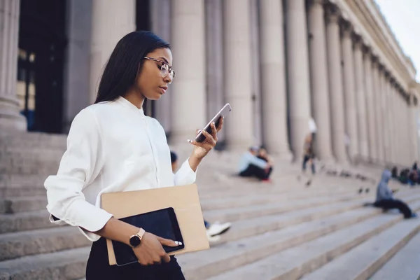 Pensive dark skinned student of faculty of law checking mail and reading notification with financial news on smartphone device connected to smartphone standing outdoors near university building