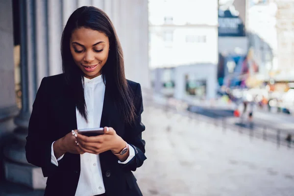 Positive african american businesswoman dressed in formal wear laughing during messaging online on smartphone device via 4G internet.Cheerful dark skinned female office worker installing application