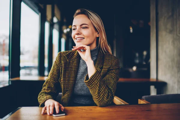 幸せな金髪の若い女はコーヒー ショップで余暇時間を過ごすを探してスタイリッシュなジャケットに身を包んだ カフェのインテリアに木製のテーブルに座って手にスマート フォンで陽気な内気な少女 — ストック写真