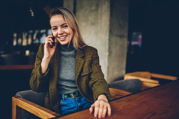Half length portrait of successful young businesswoman dressed in stylish outfit smiling at camera while talking with friend on smartphone device.Cheerful hipster blogger calling on cellular