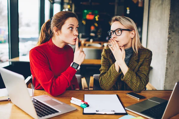 Brunette hipster blogger told secret to wondered colleague and showing sign shh during collaborating at laptop devices in coworking.Excited two best friends gossiping during studying break at netbooks