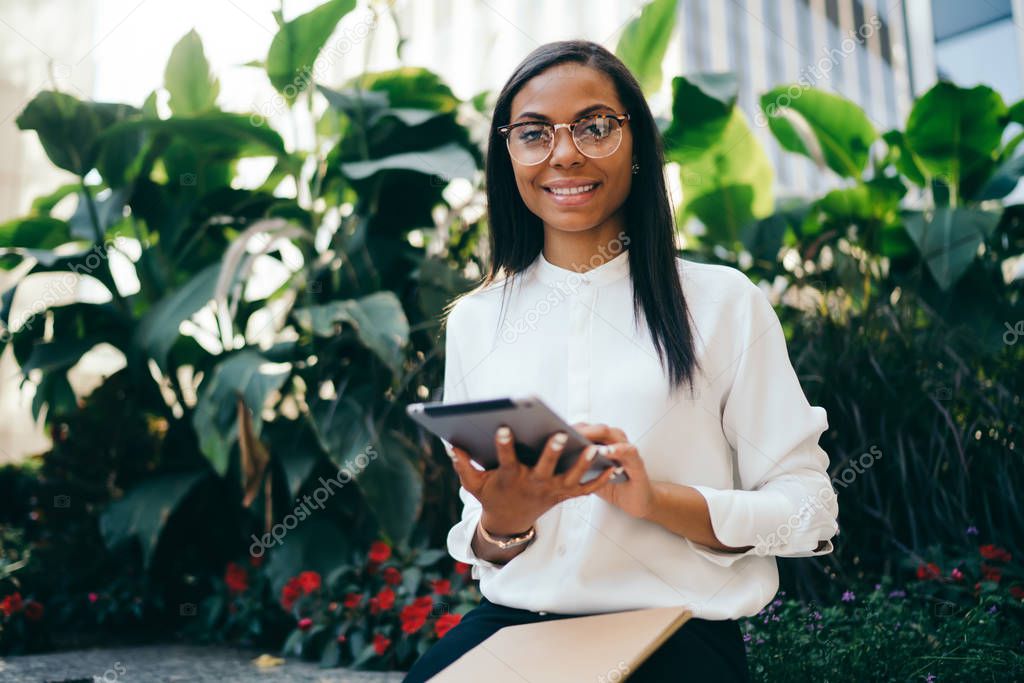 Portrait of successful dark skinned female office worker in eyewear making payment online on digital touch pad device connected to 4G internet.Prosperous african american young woman with tablet