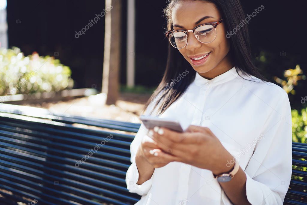 Positive african american young woman in eyewear laughing during chatting in social networks on modern smartphone.Cheerful female blogging on telephone with 4G internet sitting on wooden bench