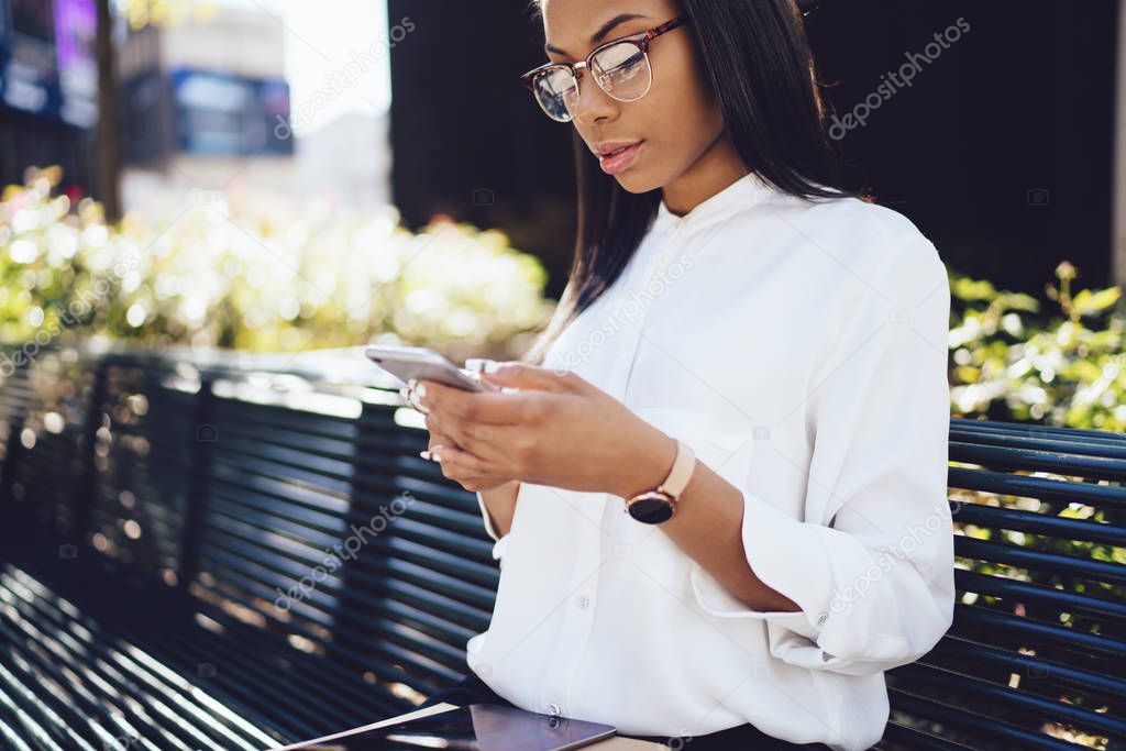 Cropped image of pensive african american student dressed in white shirt watching video on smartphone via 4G internet resting outdoors on bench.Dark skinned young woman reading news on telephone