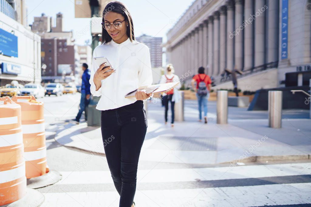 Cheerful african american female office employee dressed in elegant wear laughing during chatting online on modern smartphone device connected to 4G internet strolling outdoors in downtown