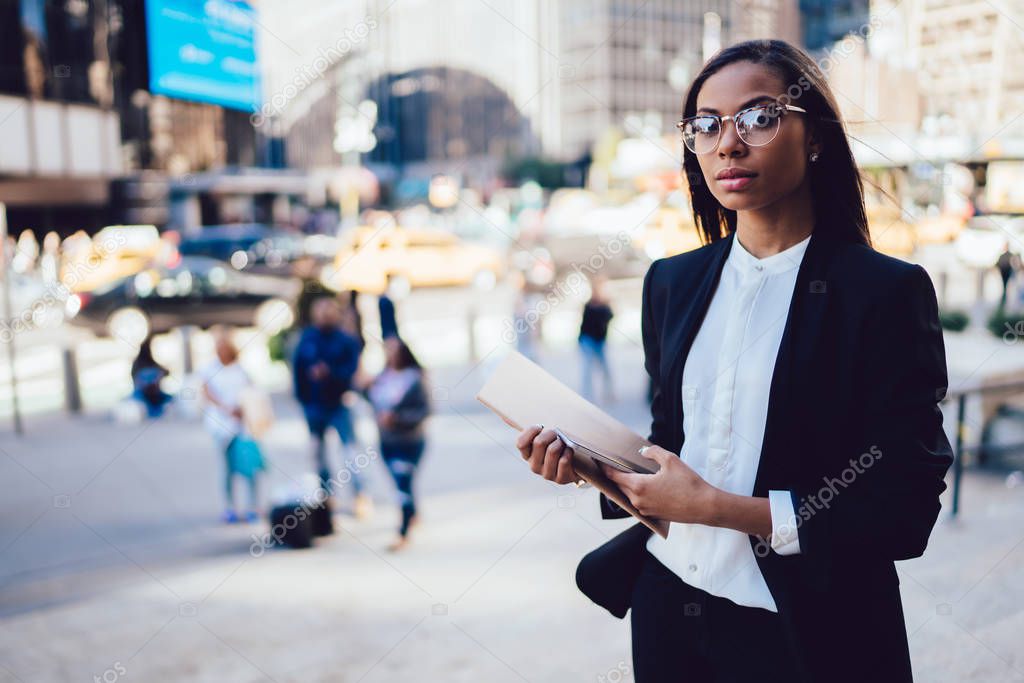 Half length portrait of african american confident businesswoman dressed in stylish formal wear holding folder and smartphone in hands while standing in downtown.Dark skinned lawyer looking at camera