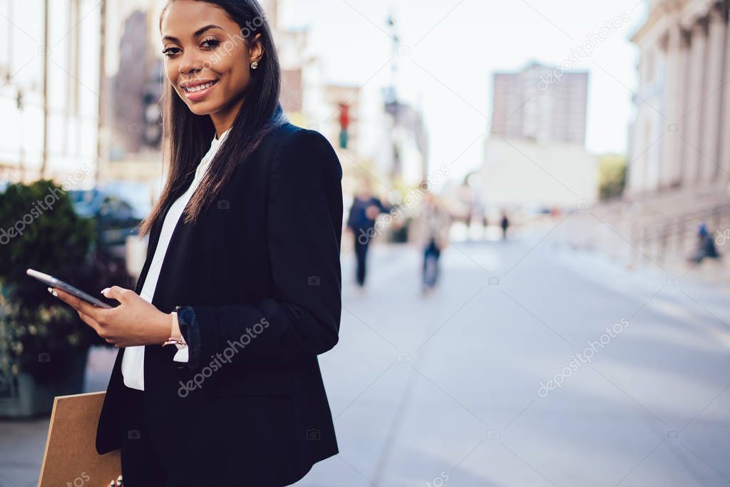 Portrait of cheerful african american office worker dressed in formal wear smiling at camera near copy space area.Successful dark skinned lawyer updating profile on smartphone standing in downtown
