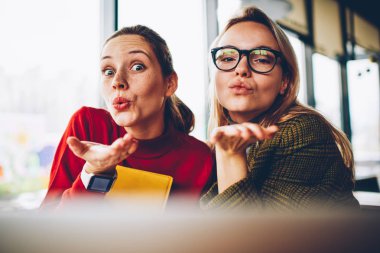 Yarım uzunluk portre neşeli genç kadınların zamana Cafe iç oturma sırasında kameraya hava öpücük gönderme rahat kıyafet giymiş. Poz ve having fun olumlu ve büyüleyici iki arkadaşları
