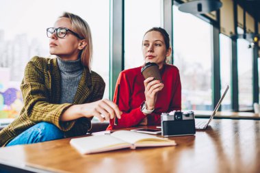 Düşünceli iki yetenekli öğrenciler giyinmiş şık casual kıyafet modern dizüstü bilgisayar, ortak proje üzerinde işbirliği için kablosuz 4 g bağlıyken pencereden dışarı bakıyor coworking internet