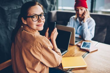 Gözlük vizyon düzeltme posta Smartphone cep telefonu ile coworking ve birlikte eğitim hipster arkadaşıyla oturan ağlarda denetlerken kameraya gülümseyen neşeli genç kadın portresi