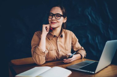 Yarım uzunluk portre ise smartphone elinde tutan ve uzaktan kablosuz internet'e bağlı dijital dizüstü bilgisayarda çalışan kameraya gülümseyen gözlük mutlu kadın grafik tasarımcısının