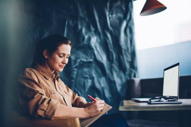 Yaratıcı öğrenci alan coworking içinde boş perde ile modern dizüstü cihaz oturan defter metin bilgileri yazma. Dalgın kadın blogger fikirler netbook çalışma Not Defteri'nde belirterek