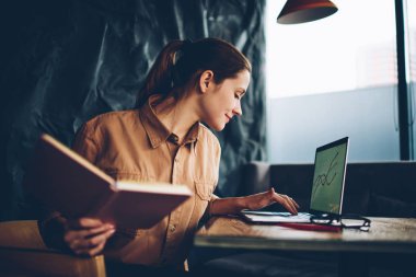 Dalgın yetenekli beyaz öğrenci dizüstü bilgisayar Projesi'nde eğitim üzerinde çalışmak için kablosuz 4 g bağlıyken ders kitabı elinde tutan Internet. Bilgi web sitelerinde arama akıllı hippi kız