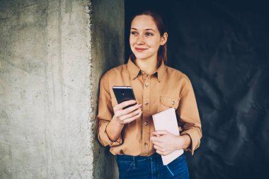 El smartphone aygıt yolu ile yüksek hızlı 4 g internet ayakta kopyalama alanı alanı yakınındaki posta kontrol kitap ile gülümseyen düşünceli genç kadın. Neşeli kadın blogger ile modern cep durulması