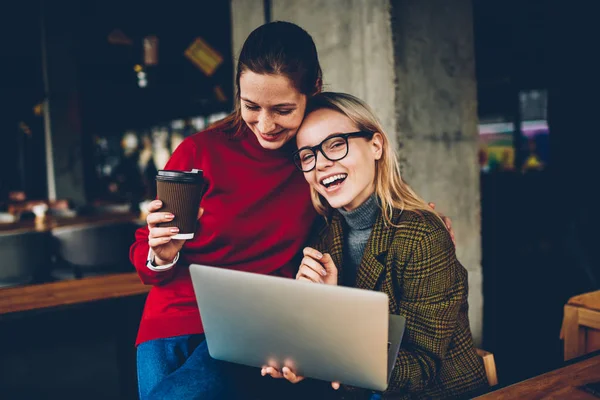 Portrait of happy blonde female blogger laughing at camera while having fun with best friend enjoying communication with each other and watching funny video on websites on laptop with 4G internet