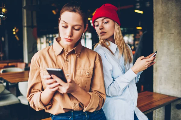 Hipster blogger spying online chat on smartphone of friend.Addicted best friends in casual outfit ignoring live communicating while spending leisure time in social networks on modern cellulars