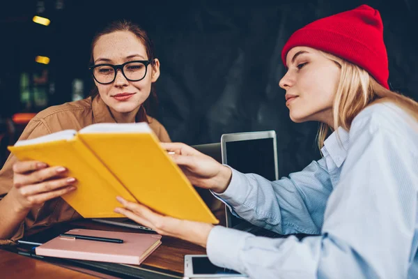 Hipster student in red hat showing textbook with useful information to colleague during collaboration on common studying task in college.Pensive smart teenagers learning educational material together