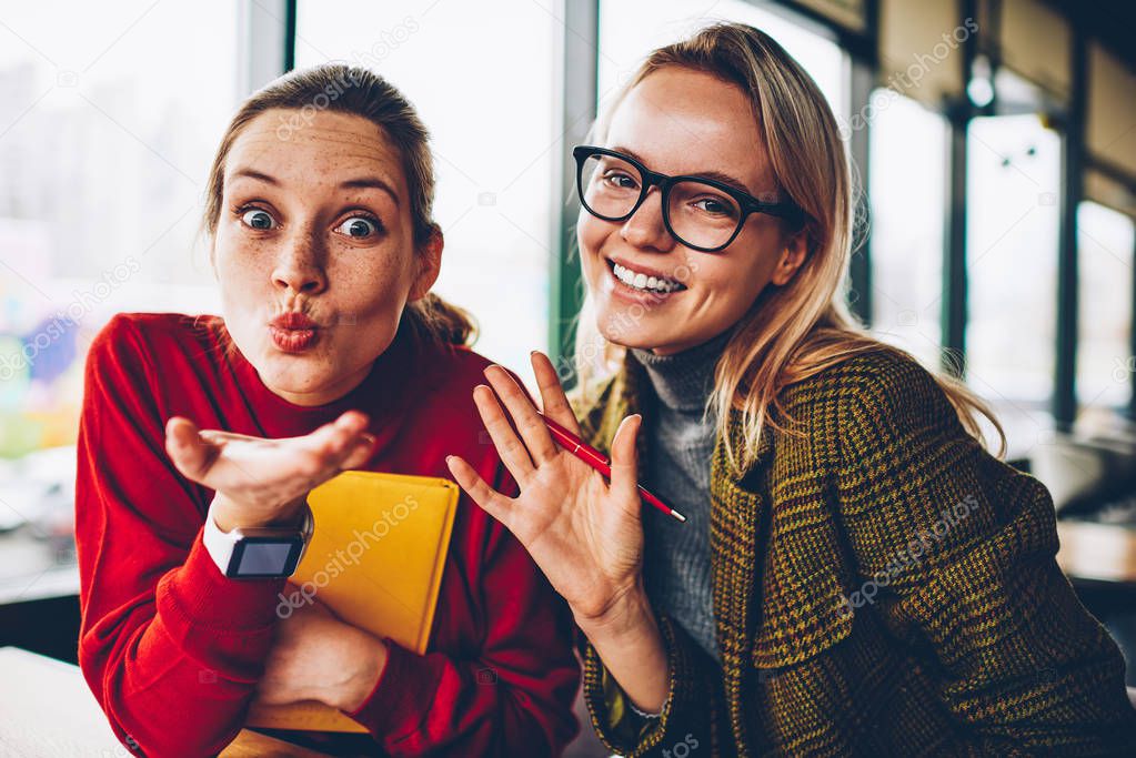 Half length portrait of positive student sending air kiss and pretty blonde hipster blogger in eyewear waving hand enjoying recreation time together.Happy young women having fun smiling at camera