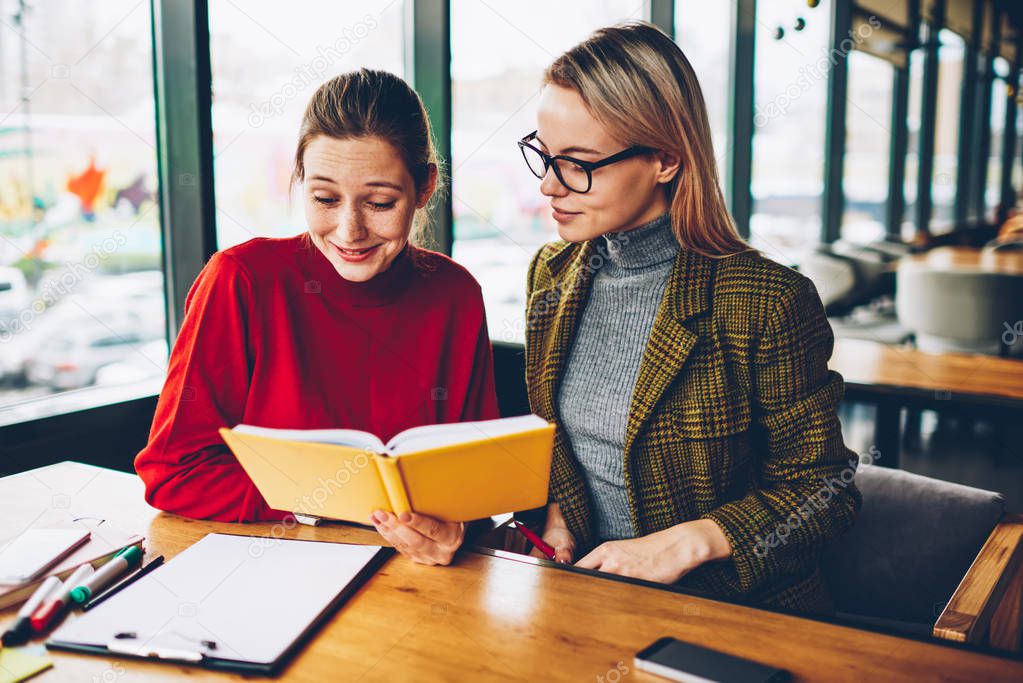 Two successful students reading textbook and studying together during exam preparation sitting in coworking.Serious female tutor with positive hipster girl conducting private lesson with literature