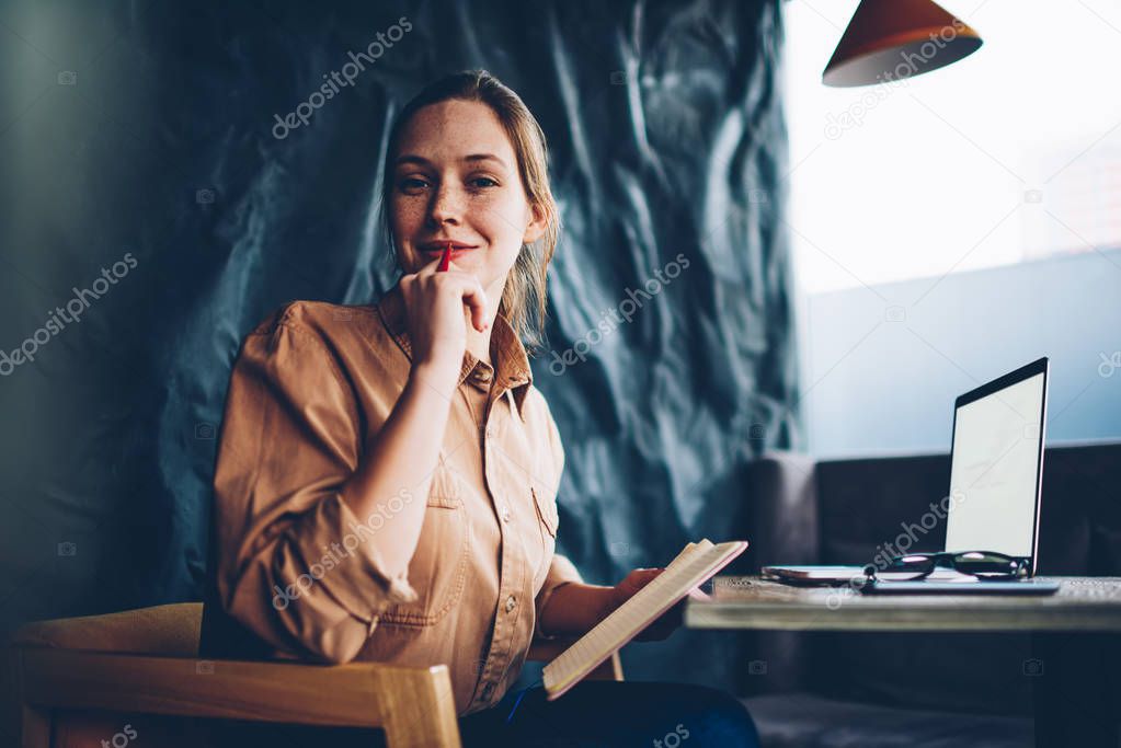 Portrait of pretty talented hipster girl with pen in hand looking at camera while sitting with notebook for notes and modern laptop computer with blank screen for productive work in coworking space