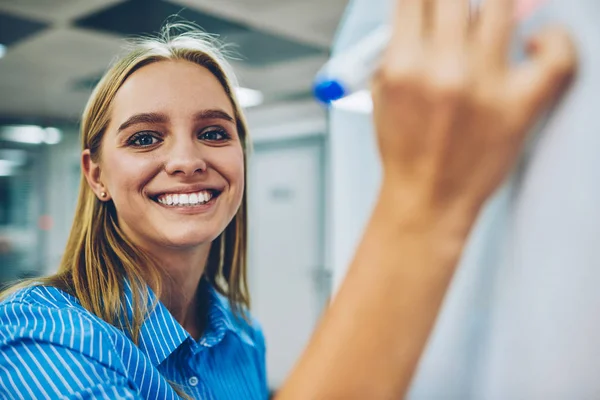 Half length portrait of cheerful blonde student writing on flipchart information for learning, creative female designer looking at camera making note of creative ideas on board planning project