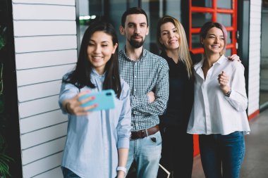 İyi görünümlü duygusal çalışan çeşitlilik grubu birlikte hücresel üzerinde çekim trendy kıyafetler giymiş. Müreffeh hipster çocuklar akıllı telefon kamera Office dururken selfie yapma