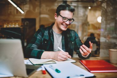 Ödev, modern teknoloji kullanılarak kafe iç çalışma olumlu müreffeh erkek serbest meslek yazarken akıllı telefon tutarak arkadaşımdan yeni iletideki gülüyor gözlük neşeli hipster adam