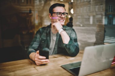 Neşeli hipster adam uzakta smartphone, müreffeh erkek freelancer mesafesi iş modern aygıtlar ve kablosuz internet ile kafeterya iç memnun tutan penceresinde arıyorum gözlükler
