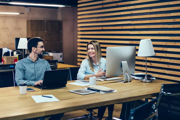 Positive male and female office employees laughing and communicating with each other during working process at computers connected to internet.Cheerful colleagues talking and discussing funny project