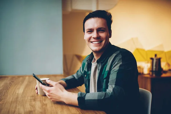 Portrait of cheerful hipster guy in casual wear enjoying coffee break chatting via telephone, happy man looking at camera smiling satisfied with good wireless connection for networking on cellphone